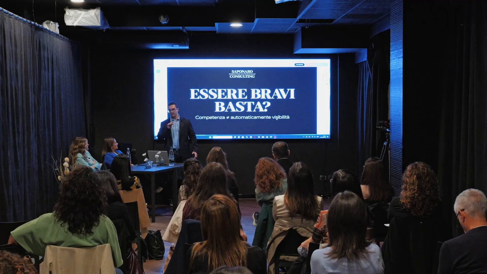 Andrea Saponaro sul palco davanti a una sala di professionisti della nutrizione, ripresa di spalle